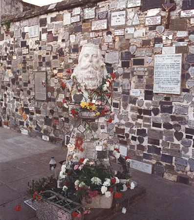 Busto de Pancho Sierra ubicado sobre un pedestal cubierto con placas, rosarios, flores e imágenes religiosas. Detrás, una de las paredes exteriores del cementerio de Salto con las placas de agradecimiento